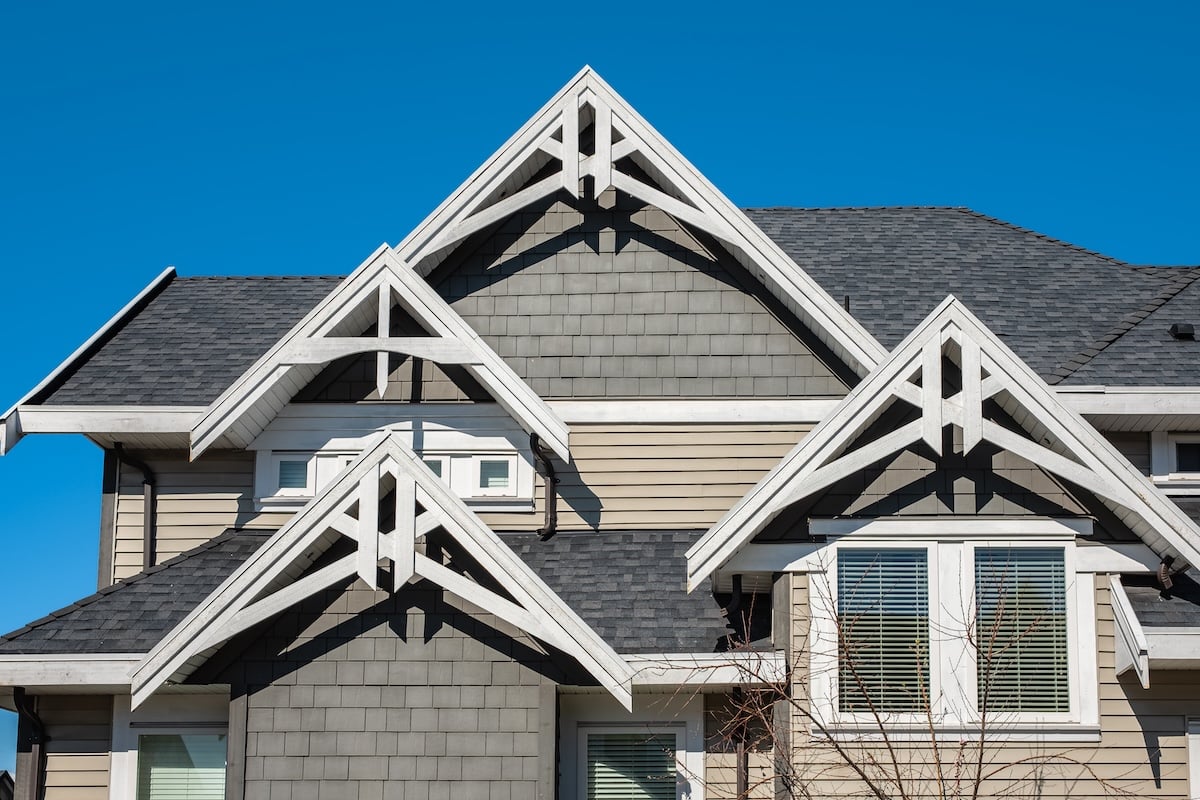 Modern house exterior with overlapping peaked gables, gray siding, white trim, and multiple windows under a clear blue sky.