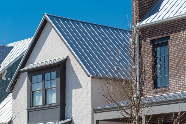 Close-up of modern houses featuring metal roofs, white and brown brick exteriors, large windows with dark frames, and a clear blue sky in the background.