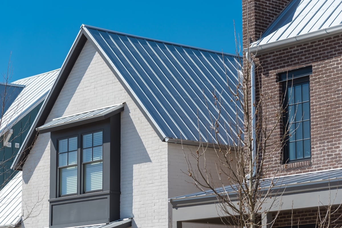 Close-up of modern houses featuring metal roofs, white and brown brick exteriors, large windows with dark frames, and a clear blue sky in the background.