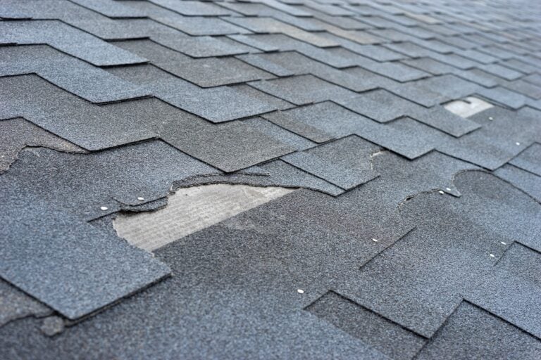 A close-up view of damaged asphalt roof shingles, with some shingles missing or torn, exposing the underlayment beneath.
