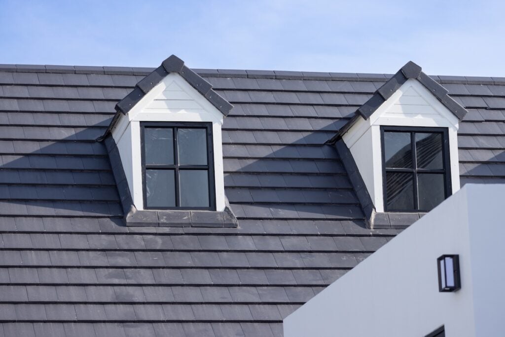 Energy Efficient Roofing Virginia A close-up view of a modern grey tiled roof with two white dormer windows, each featuring black window frames, under a clear blue sky.