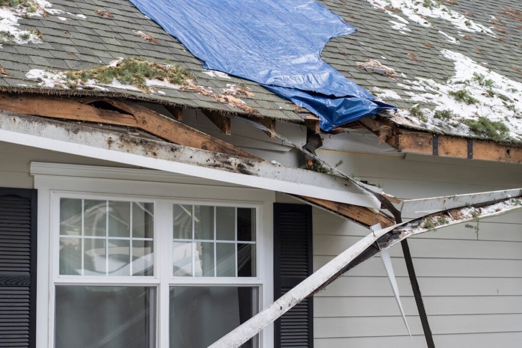 Repair Or Replace Roof Alexandria VA A house with roof damage covered by a blue tarp. The roof is missing shingles, with exposed wood and some snow on top. Broken gutters hang loosely above a white window with black shutters.