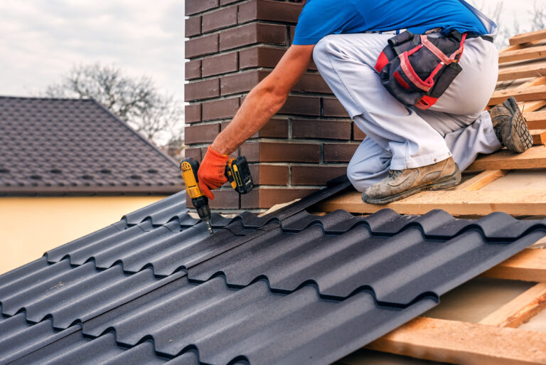 A worker in gloves uses a drill to install black metal roofing sheets around a brick chimney on a wooden roof frame.