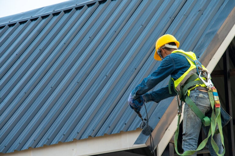 A construction worker in a safety harness, yellow helmet, and reflective vest installs or repairs a dark metal roof on a building. The worker is using tools and standing on scaffolding near the roof’s edge.