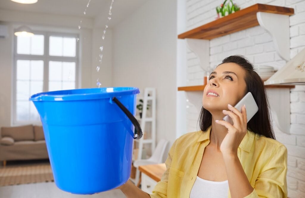 Common Roof Problems Alexandria VA A woman holds a blue bucket to catch water dripping from the ceiling—one of the common roof problems Alexandria VA residents face—as she talks on the phone in a bright, modern living room.