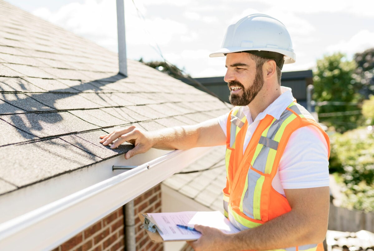 A man wearing a white hard hat and orange safety vest inspects roof shingles on a house, clipboard in hand, highlighting the importance of regular Roof Inspection Frequency in Alexandria while standing next to a white gutter in sunlight.