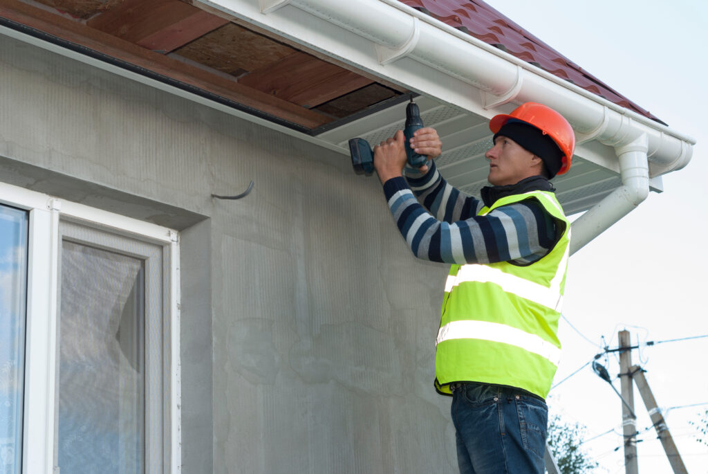 Roof Repair Cost Alexandria VA A construction worker in a neon safety vest and orange helmet uses a power drill to work on the soffit under the eaves of a house with gray walls and a red roof.