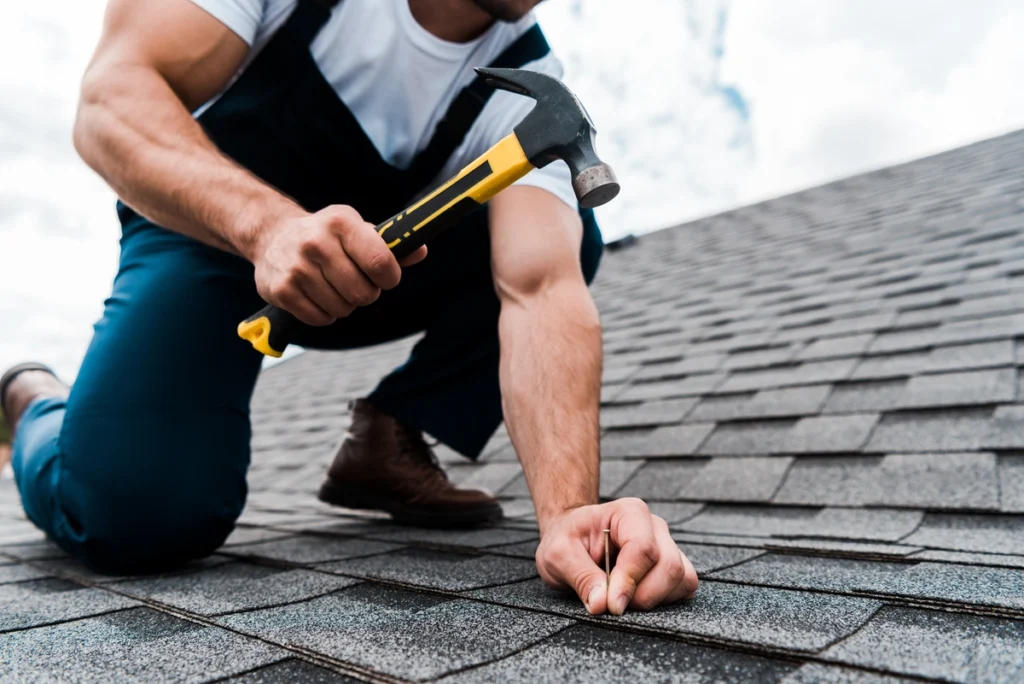 A person in work clothes kneels on a shingled roof, using a hammer and holding a nail, likely repairing or installing shingles under a partly cloudy sky.