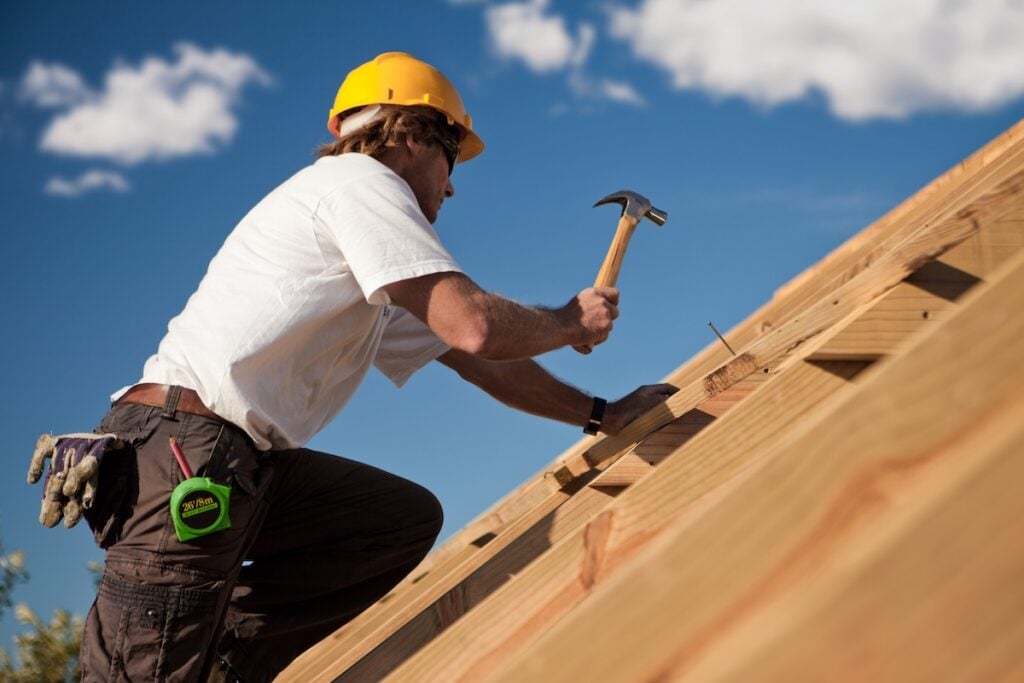 A construction Best Roofing Companies Alexandria VA worker in a yellow hard hat and white shirt uses a hammer to drive a nail into a wooden roof frame under a blue sky with clouds. A tape measure hangs from his belt.