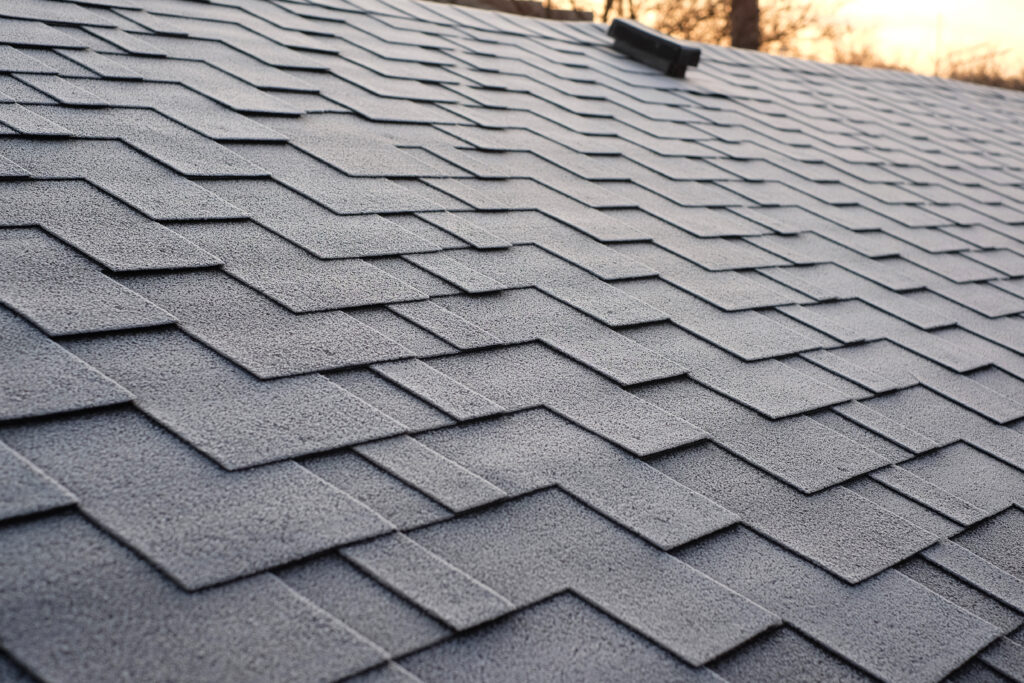 Roof Repair Cost Alexandria VA Close-up view of gray asphalt shingles arranged in a staggered pattern on a sloped roof, with early morning or evening sunlight casting a warm glow in the background.