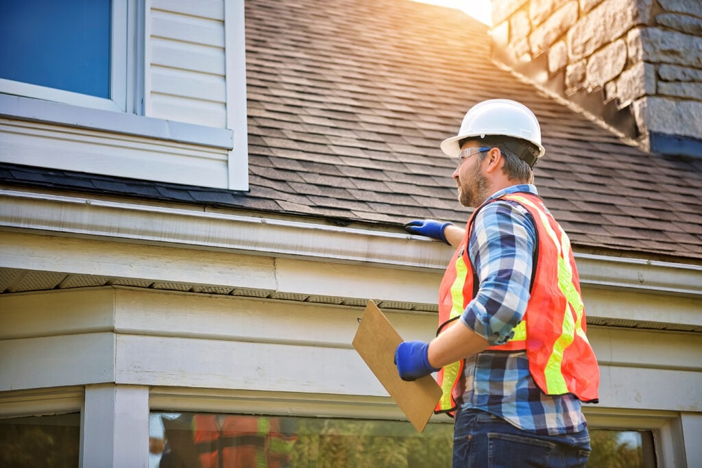 Roof Inspection Frequency Alexandria A construction worker in a white hard hat, safety vest, and gloves inspects the roof of a house, holding a clipboard. The worker checks the shingles near the edge on a sunny day, ensuring proper roof inspection frequency in Alexandria.