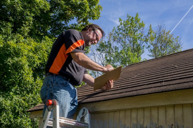 A man in safety glasses stands on a ladder next to a house, inspecting the roof and writing on a clipboard, surrounded by green trees under a blue sky.
