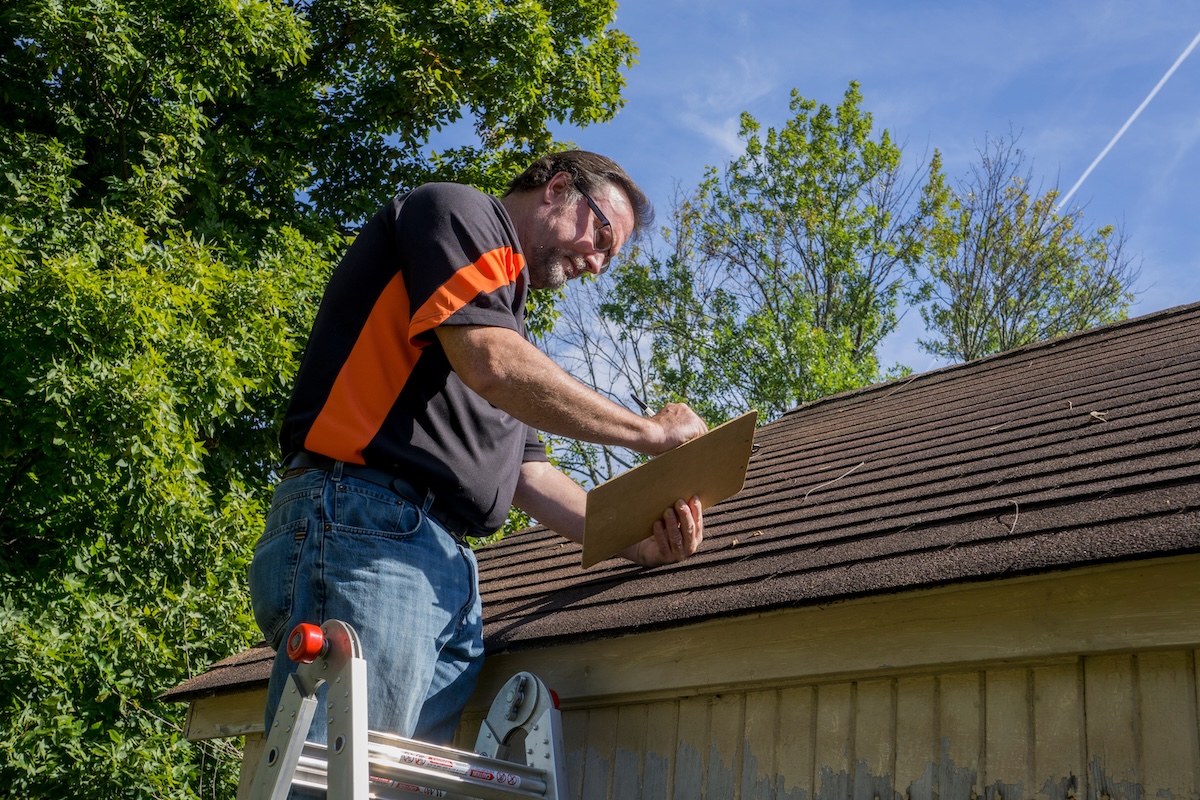 A man in safety glasses stands on a ladder next to a house, inspecting the roof and writing on a clipboard, surrounded by green trees under a blue sky.