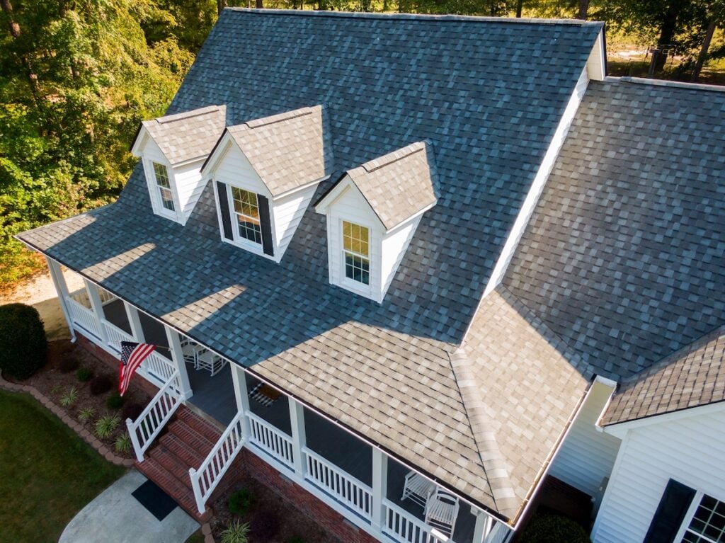 Roof Inspection Frequency Alexandria Aerial view of a house with gray shingle roofing, white siding, dormer windows, and a large covered front porch. An American flag hangs near the entrance. Trees and lawn surround the home—ideal for discussing roof inspection frequency in Alexandria.