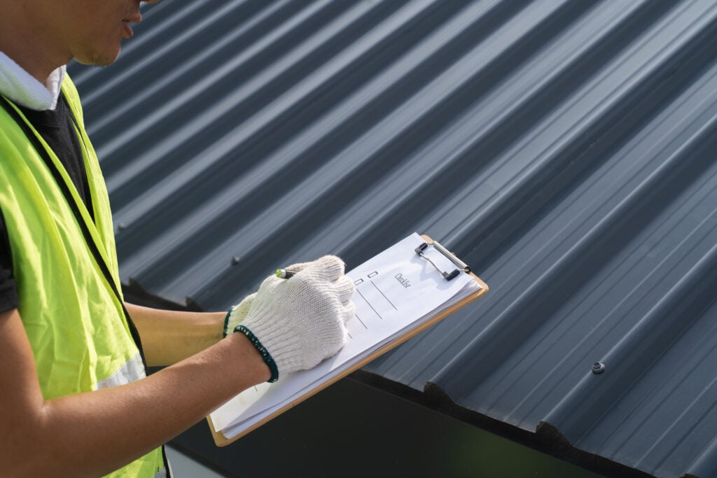 Roof Inspection Frequency Alexandria A person wearing a yellow safety vest and white gloves holds a clipboard and writes notes while inspecting a dark metal roof, demonstrating the importance of regular Roof Inspection Frequency in Alexandria. The person's face is partially out of frame.