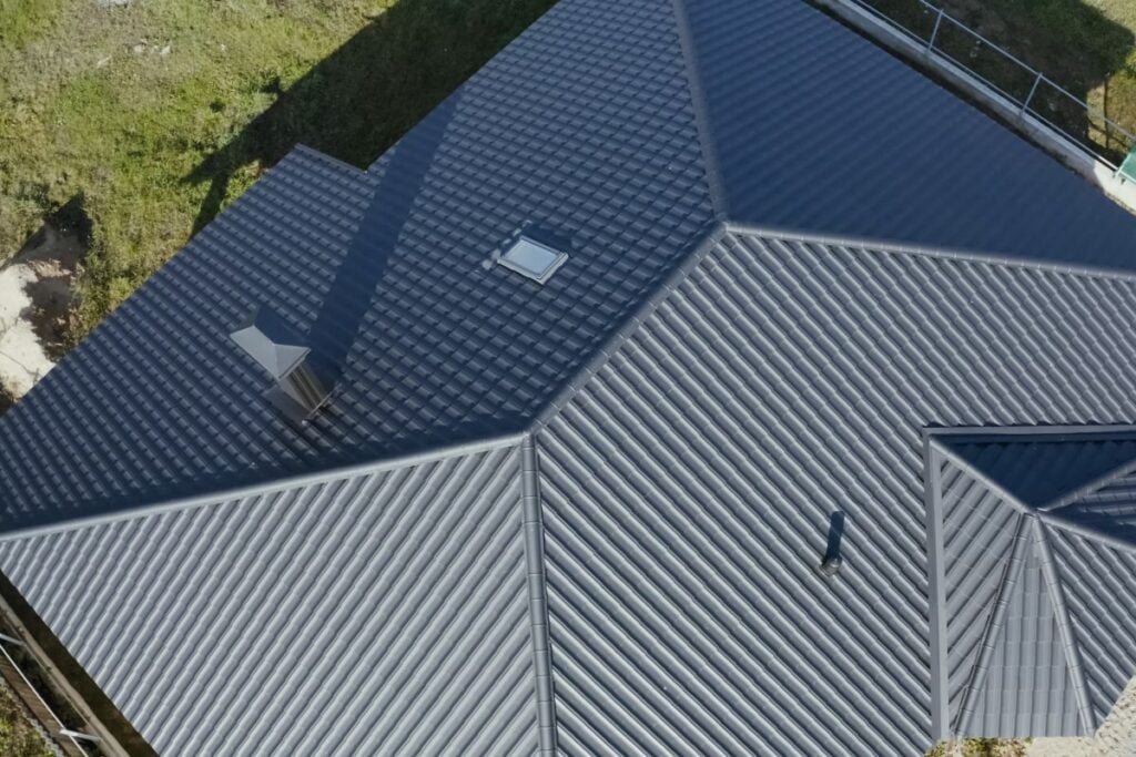 Roofing Companies In Fairfax VA Aerial view of a modern house roof with dark gray metal panels, featuring a small skylight, chimney, and surrounding green grass.