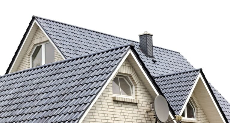 A close-up of a house roof with gray metal roof tiles, white brick walls, arched windows, a chimney, and a satellite dish, all set against a white background.