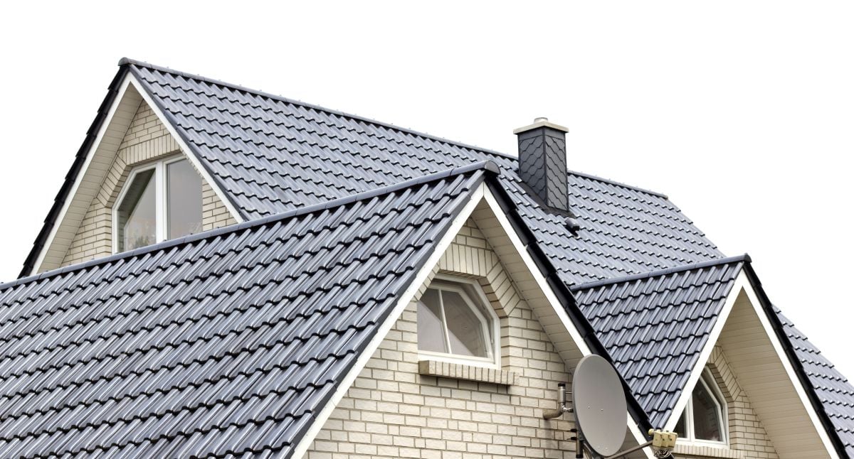 A close-up of a house roof with gray metal roof tiles, white brick walls, arched windows, a chimney, and a satellite dish, all set against a white background.