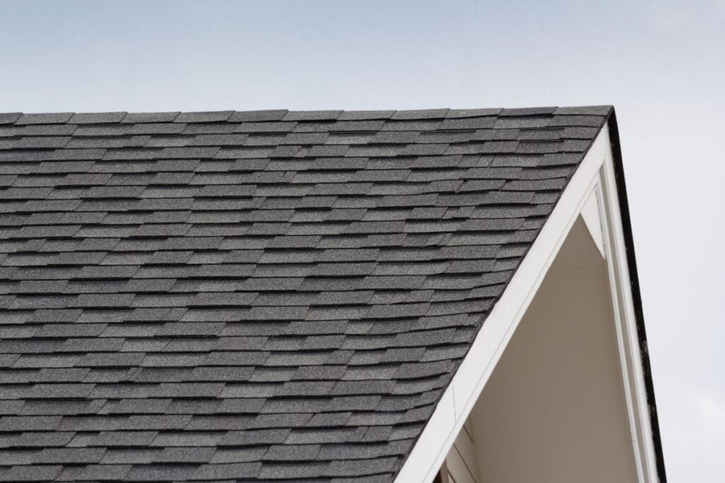 Close-up view of a house roof with dark gray asphalt shingles and white trim against a pale blue sky. The neatly arranged shingles showcase the quality work often delivered by top roofing companies in Burke VA.