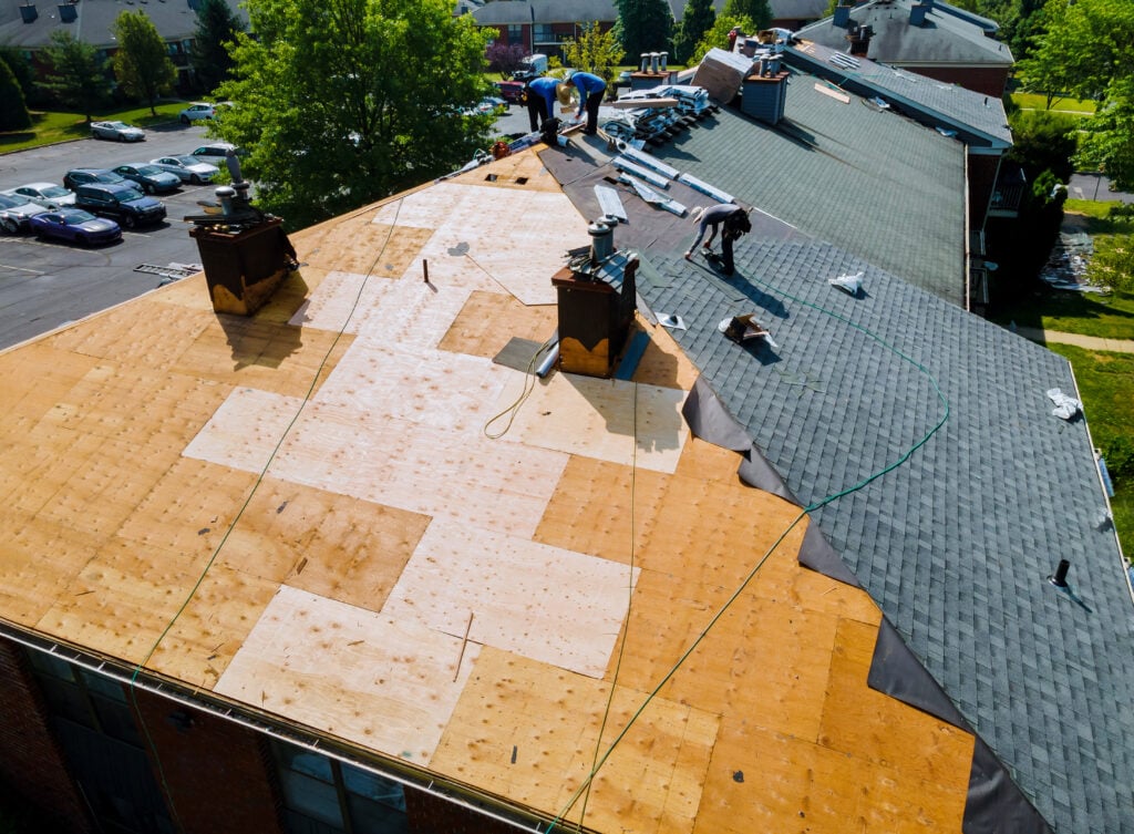 Aerial view of workers replacing shingles on a large residential roof, with sections covered in plywood and some areas still under construction. Tools and materials are scattered across the rooftop.