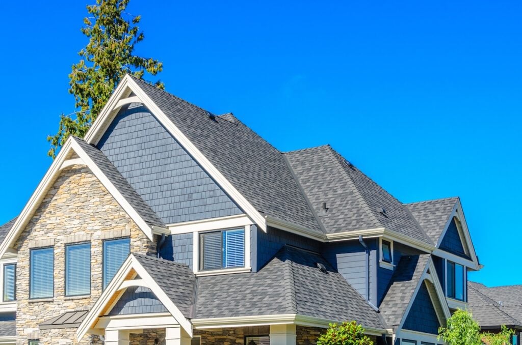 Modern suburban house with gray shingles, stone accents, and white trim under a clear blue sky. Multiple gables and large windows showcase quality roofing Burke is known for, all surrounded by lush greenery.