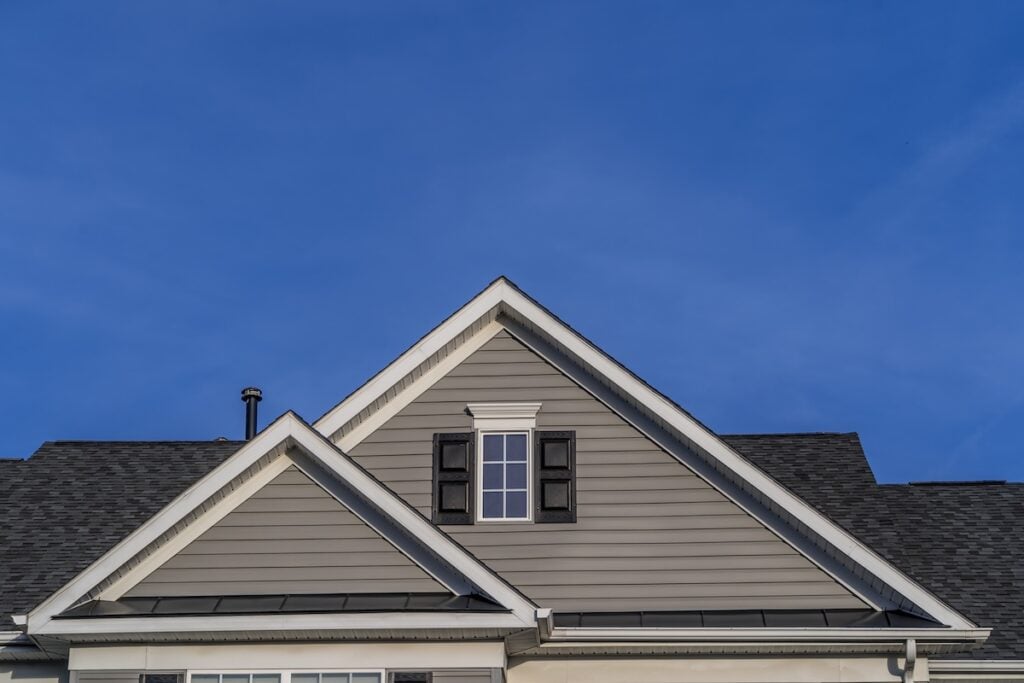 The upper portion of a house with gray siding, black roof shingles installed by roofing Arlington VA experts, a central window with black shutters, and a clear blue sky in the background.