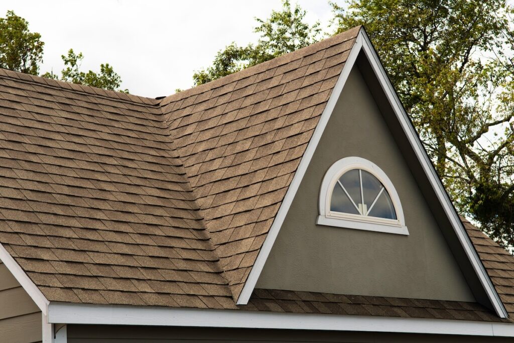 Close-up of a house roof with brown asphalt shingles installed by roofing McLean VA, featuring a small arched window. The gray siding and white trim outline the window and roof edges, with green trees visible in the background.