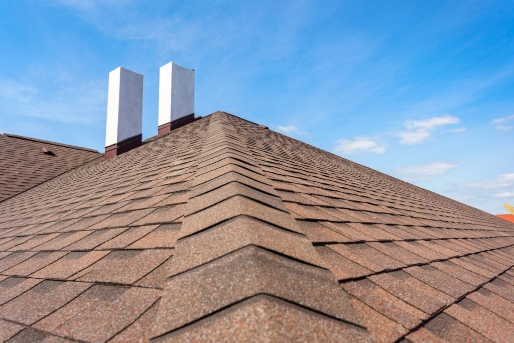 Close-up view of a brown shingle roof with two white rectangular chimneys extending above it, under a clear blue sky with a few scattered clouds—expert roofing Burke craftsmanship on display.