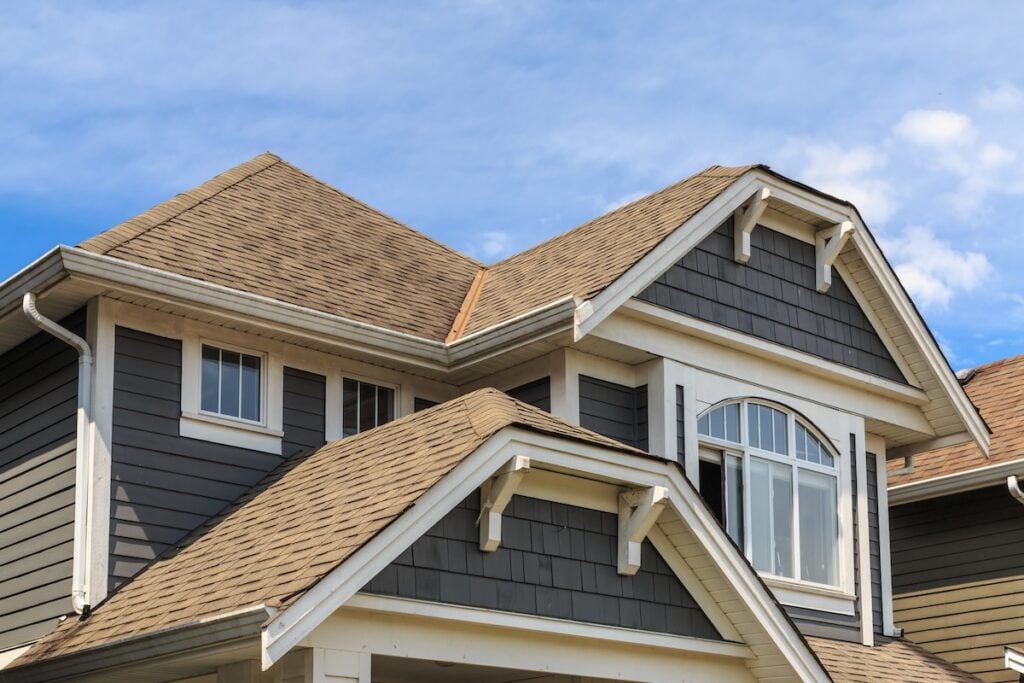 Close-up of the upper part of a modern house with dark gray siding, white trim, and multiple gabled roofs, showcasing expert roofing McLean VA craftsmanship against a blue sky with some clouds.