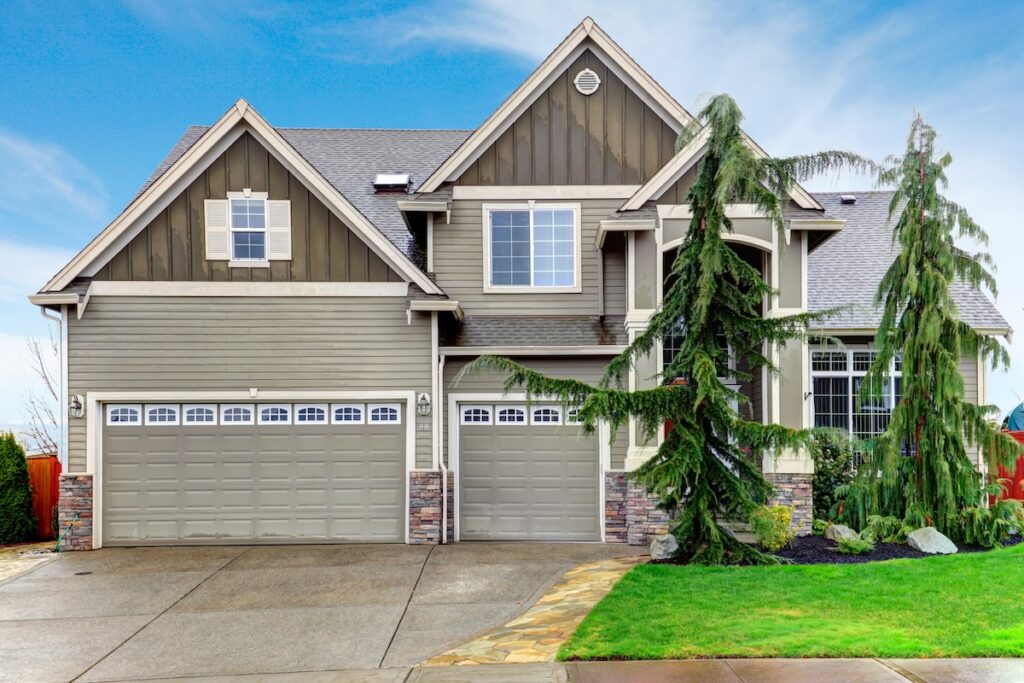 Two-story suburban house with gray siding, large windows, and three-car garage. The front yard features green grass, stone accents, a pathway, and tall evergreen trees—expertly maintained by roofing Fairfax Station professionals.