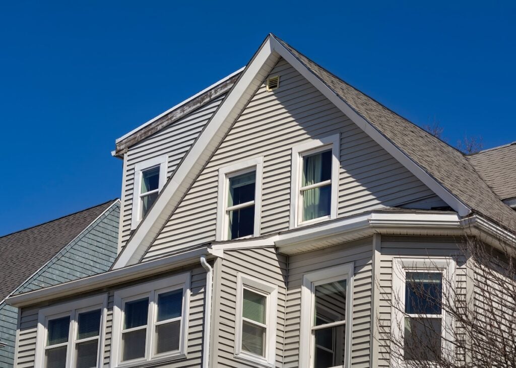 A close-up view of the upper portion of a light gray house with multiple windows, gabled roofs, and white trim showcases quality roofing Burke craftsmanship against a clear blue sky.
