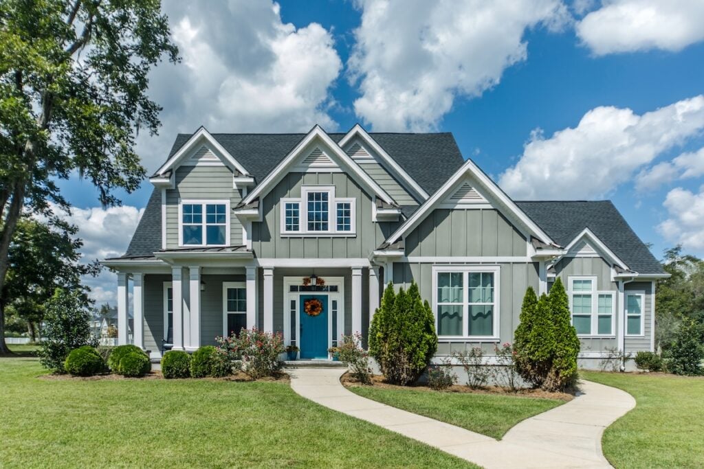 A modern two-story gray house with white trim, blue front door, columns, and gabled roof showcases expert roofing Burke style, surrounded by green lawn, shrubs, and a curved walkway under a partly cloudy sky.