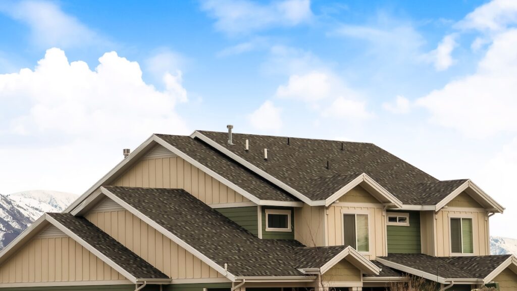 A modern two-story house with green and beige siding and multiple gabled, shingled roofs—showcasing expert roofing Fairfax Station craftsmanship—set against a bright blue sky with white clouds and distant mountains.