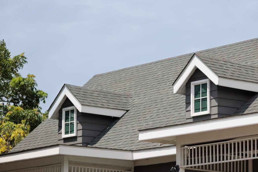The image shows the upper part of a house with a gray shingle roof installed by a Virginia roofing company, two dormer windows, white trim, and part of a veranda railing. Green trees and a blue sky are visible in the background.