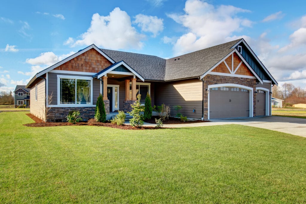 Single-story suburban house with a two-car garage, brown and beige siding, stone accents, and new roofing. Arlington VA charm shows in the front porch, large windows, and well-maintained green lawn under a blue sky with scattered clouds.