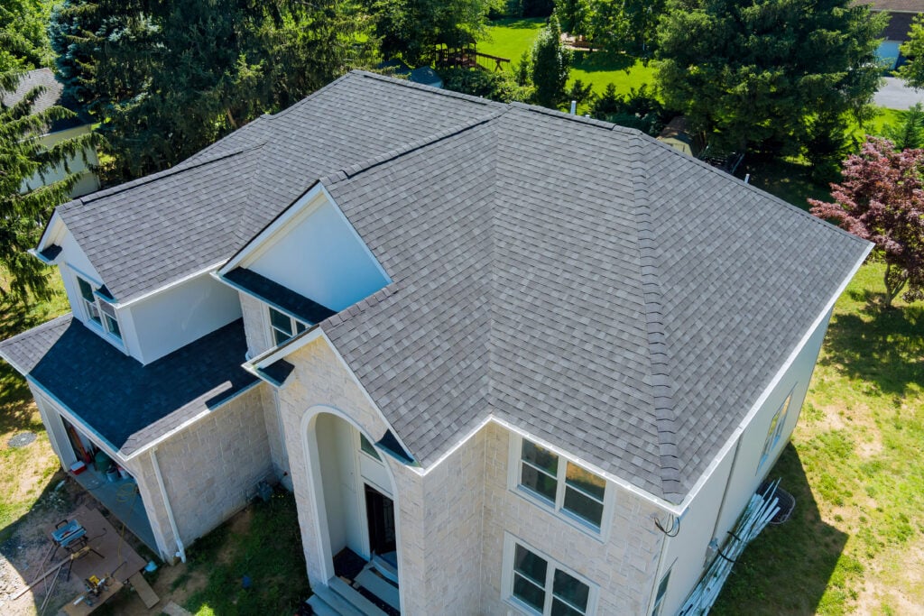 Aerial view of a large, two-story house with light-colored stone exterior and a dark gray shingled roof, expertly installed by roofing Alexandria professionals, surrounded by trees and green lawn.
