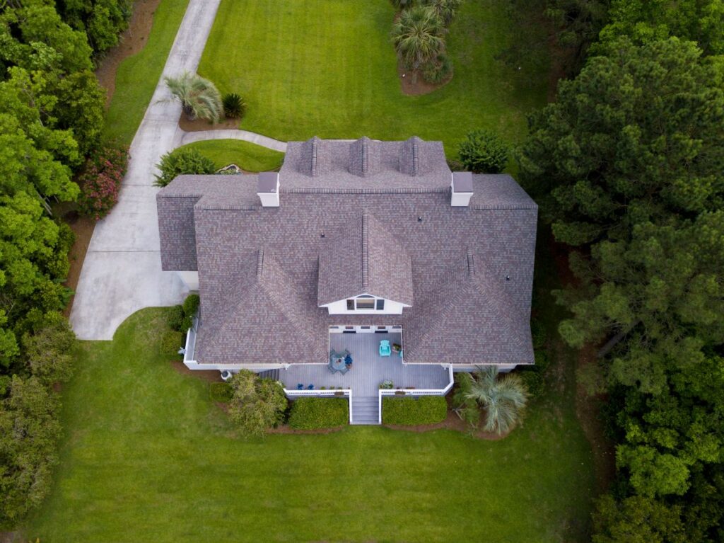 Aerial view of a large house with a gray roof, surrounded by green lawn and trees. The roofing Springfield homes trust adds curb appeal. A driveway curves left to the back, and outdoor seating is visible on the front porch.