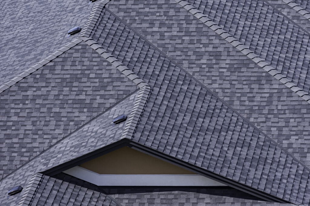 Close-up view of intersecting gray shingled roof sections by Roofing Alexandria, forming geometric patterns with sharp angles and overlapping lines. The uniformly arranged shingles and visible attic vents enhance the roof's visual appeal.