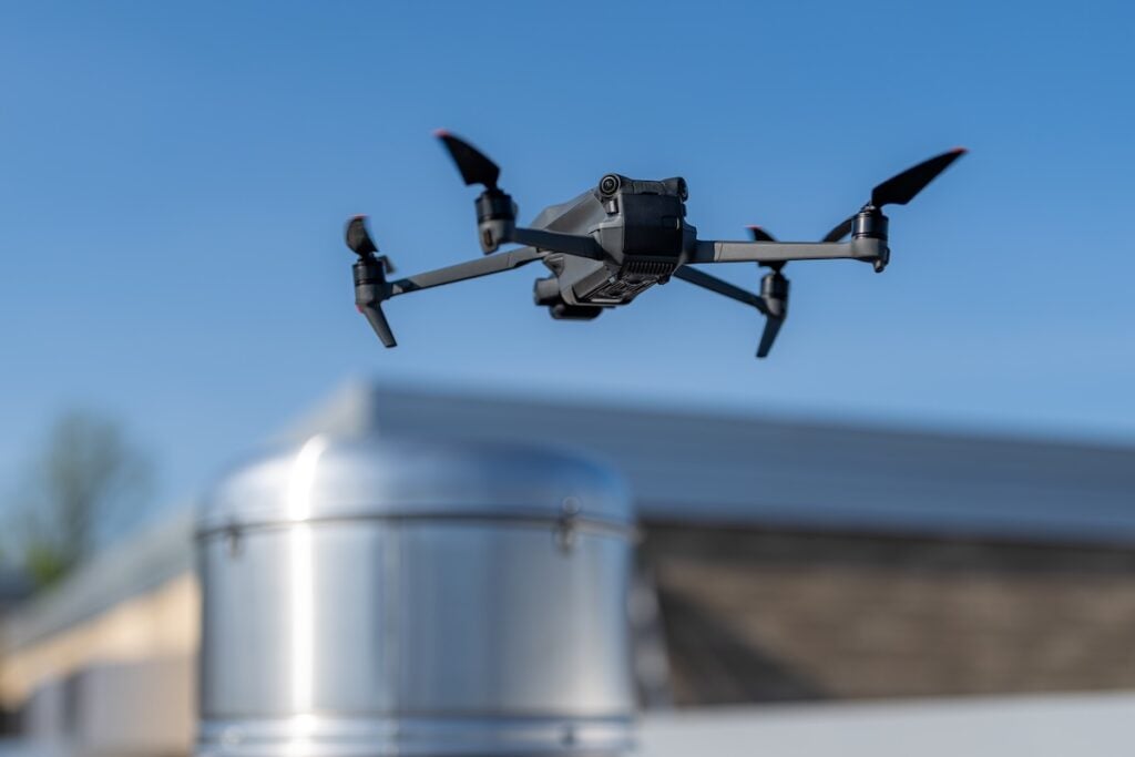 A black quadcopter drone flying outdoors near a metallic cylindrical structure, with a blurred building and blue sky in the background—perfect for capturing roofing Alexandria projects from above.