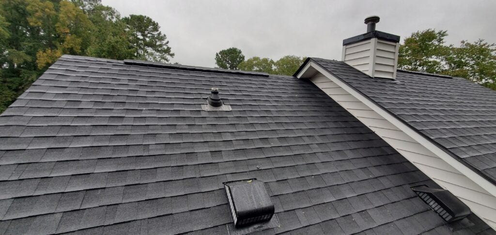 Gray shingled house roof with two vent pipes and a roof vent, next to a white chimney with black trim. Roofing Arlington VA experts ensure durability, with trees and a cloudy sky visible in the background.