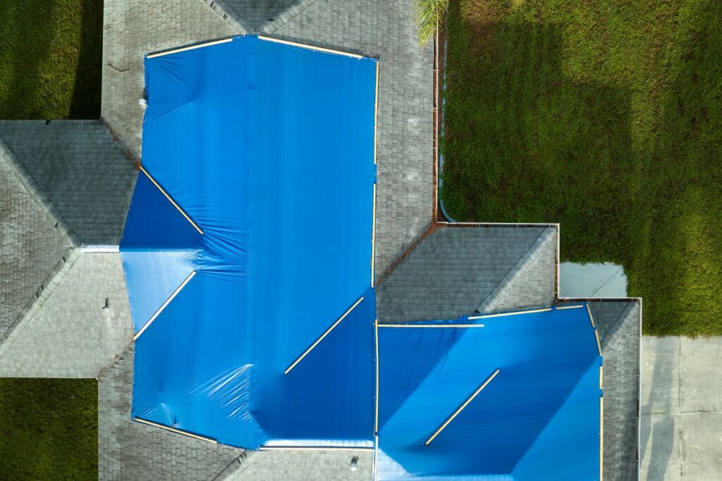 Aerial view of a house roof covered with large blue tarps, weighed down by wooden planks, near green grass and a concrete driveway—an example of emergency roofing in Mount Vernon.