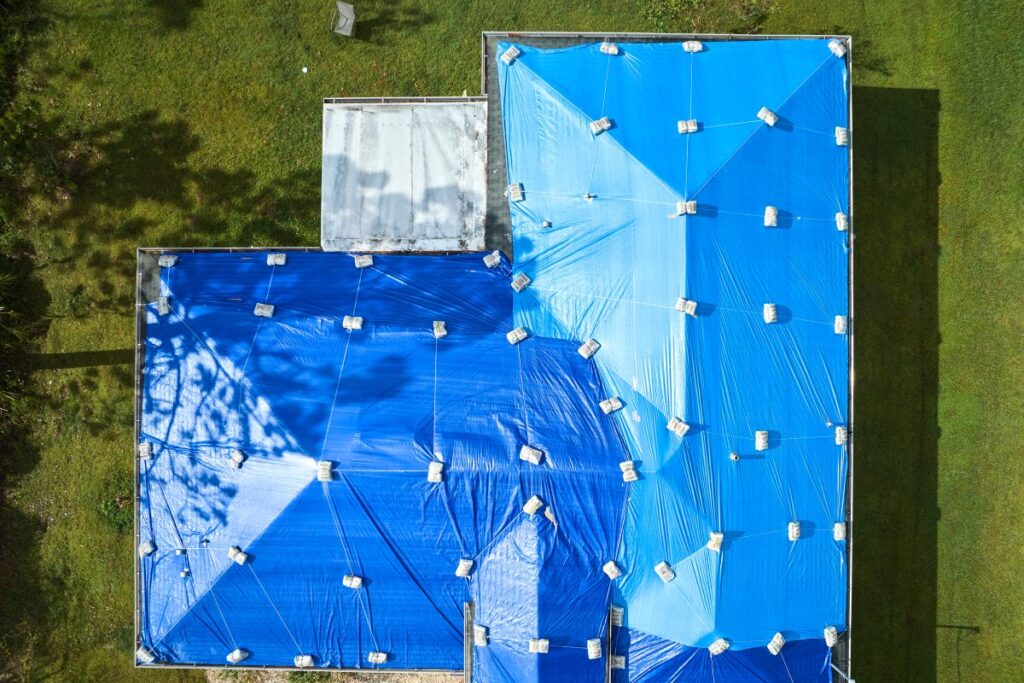 Aerial view of a house roof covered with blue tarps secured by sandbags, surrounded by green grass. The tarps suggest ongoing roof repair in Lorton, likely protecting the home from further damage.