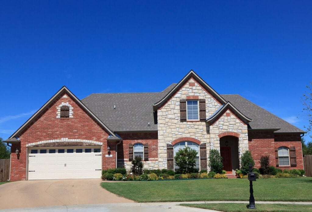 A large suburban house with a mix of red brick and light stone exterior, gray roof by roofing Arlington VA experts, a two-car garage, green lawn, small shrubs, and a black mailbox on a sunny day with a clear blue sky.