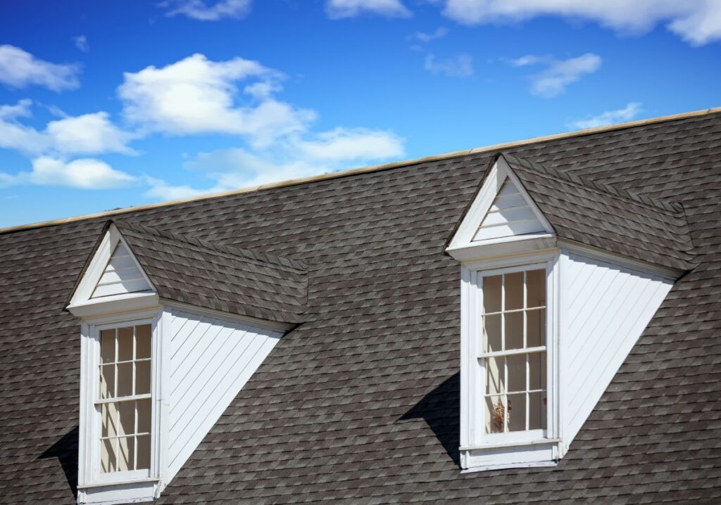 Two white dormer windows with triangular roofs on a gray shingled roof, expertly crafted by Roofing Mount Vernon, set against a blue sky with scattered white clouds.