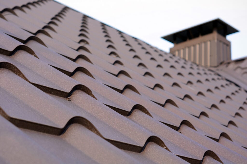 Close-up view of a brown metal tile roof with a vented chimney structure in the background, highlighting the wavy pattern and texture of the roofing material—an example of expert roofing McLean VA craftsmanship.