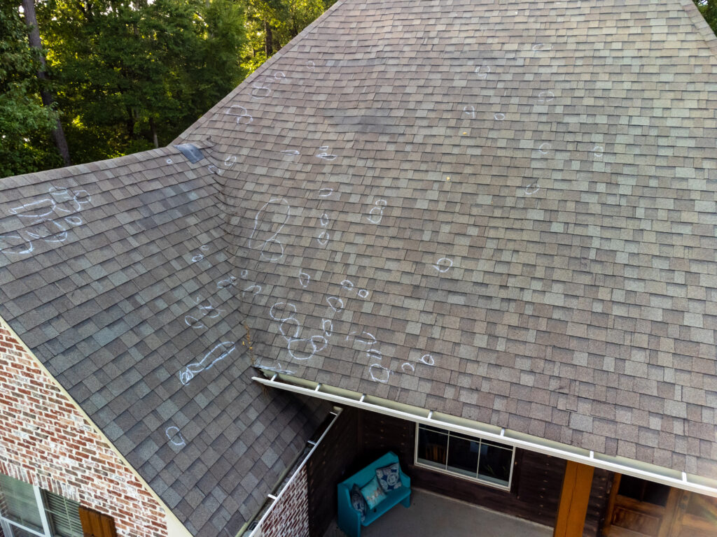 Aerial view of a house roof with multiple chalk circles and marks highlighting hail damage on the shingles. A small patio with a blue bench is visible below. Trees surround the house.