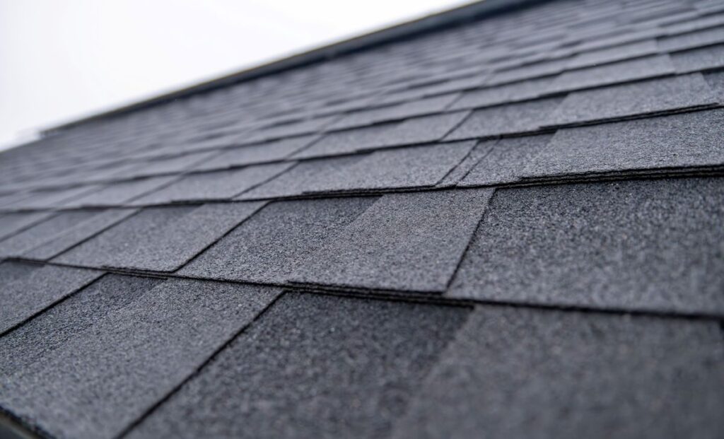 Close-up view of dark gray asphalt roof shingles, showing their textured surface and overlapping arrangement. The image captures the angled slope of a roofing Mount Vernon project, leading toward a bright, blurred sky.
