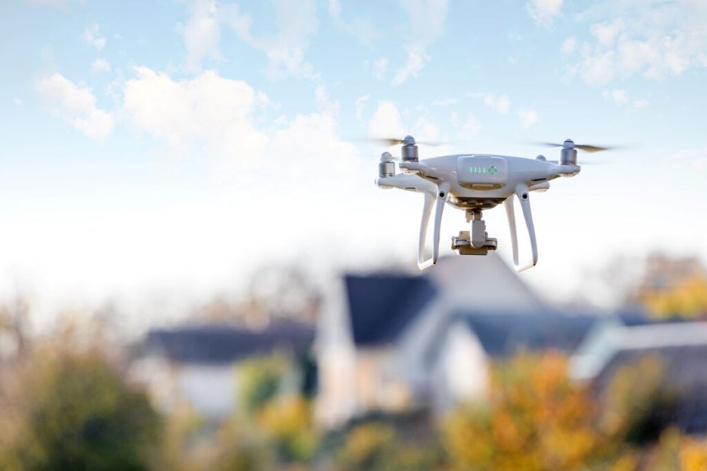 A white quadcopter drone hovers outdoors, surveying rooftops with a blurred background of trees and houses in Mount Vernon, perfect for roofing inspections under a blue sky with clouds.