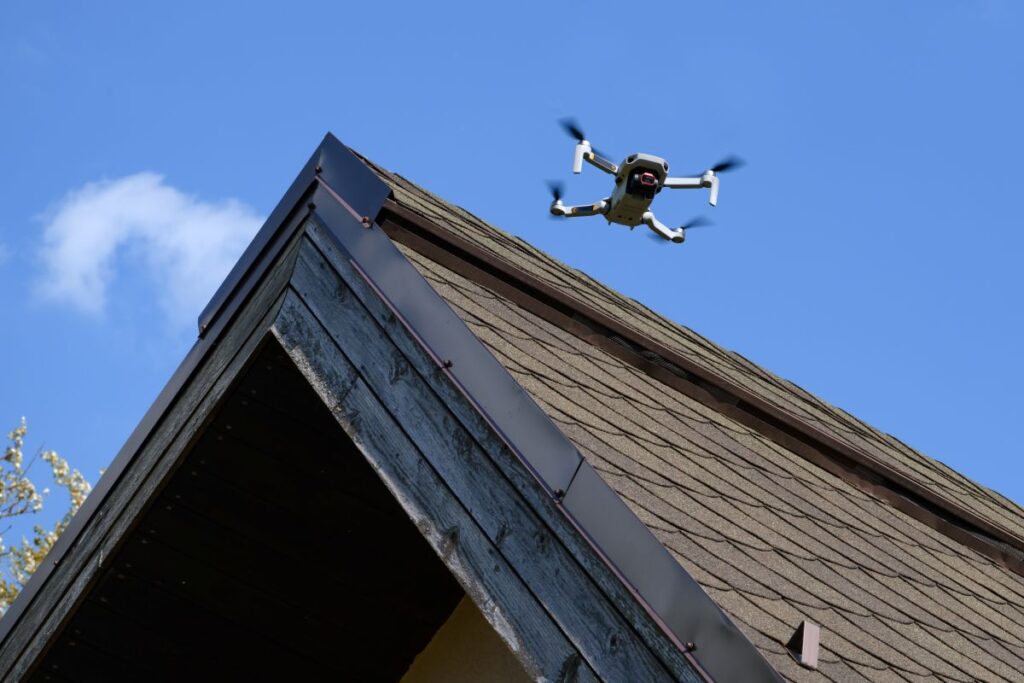 A small drone with four propellers flies above the roof of a wooden house, capturing images for a roofing Springfield inspection against a clear blue sky.