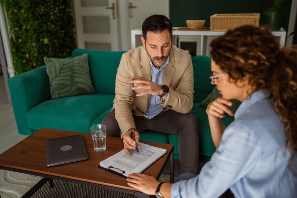 Two people sit on chairs near a coffee table, discussing roof repair in Lorton with notes. One gestures while speaking, the other listens thoughtfully. A laptop and glass of water are on the table, with a green sofa in the background.